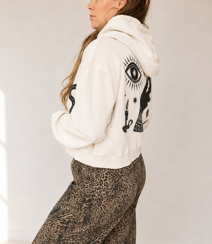 A woman wearing a cream-colored hoodie with black graphic prints on the back and wide-leg leopard-print pants, standing in a brightly lit room.