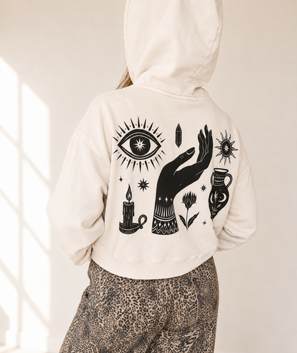 A person shown from the back wearing a cream-colored hoodie with a large black graphic print of a hand, eye, candles, and flowers, paired with leopard-print pants in a bright indoor setting.
