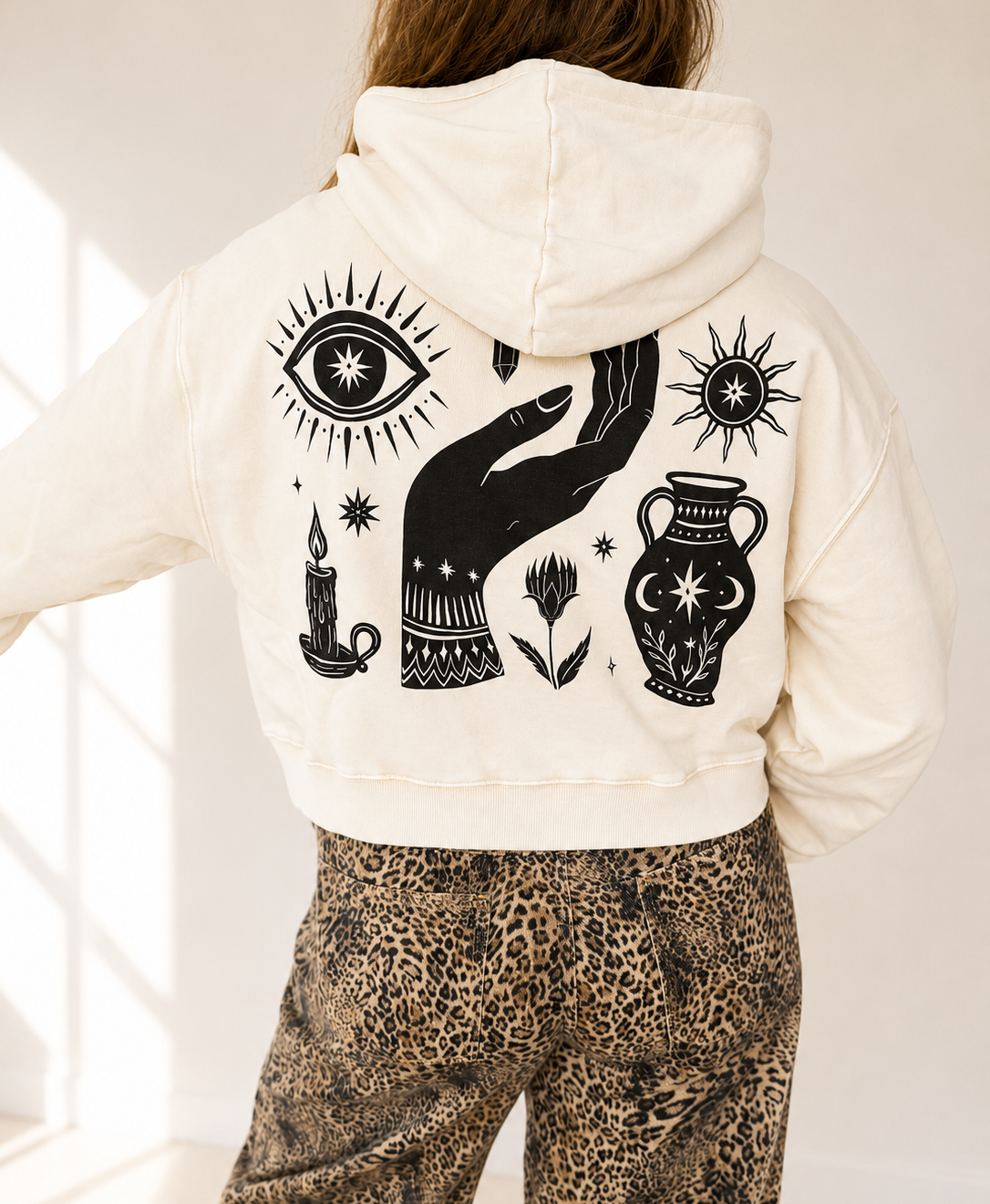 A person shown from the back wearing a cream hooded sweatshirt with a large black graphic print featuring an eye, hand, candle, flower, urn, and sun, paired with leopard-print pants in a bright indoor setting.
