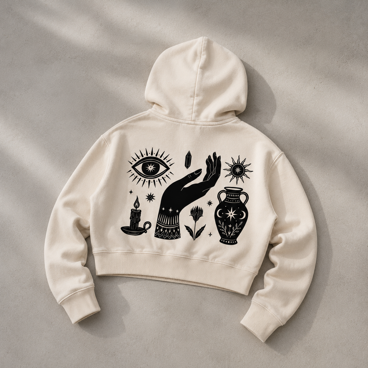 An off-white cropped hoodie with a black graphic print of a hand, eye, candle, flower, and urn, laid flat on a textured gray background.