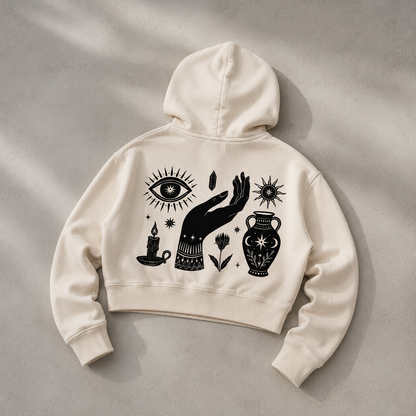 An off-white cropped hoodie with a black graphic print of a hand, eye, candle, flower, and urn, laid flat on a textured gray background.