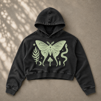 A black washed cropped hoodie with a large light green graphic of a moth, snake, leaf, and moon on the front, laid flat on a textured beige surface with dappled shadows.