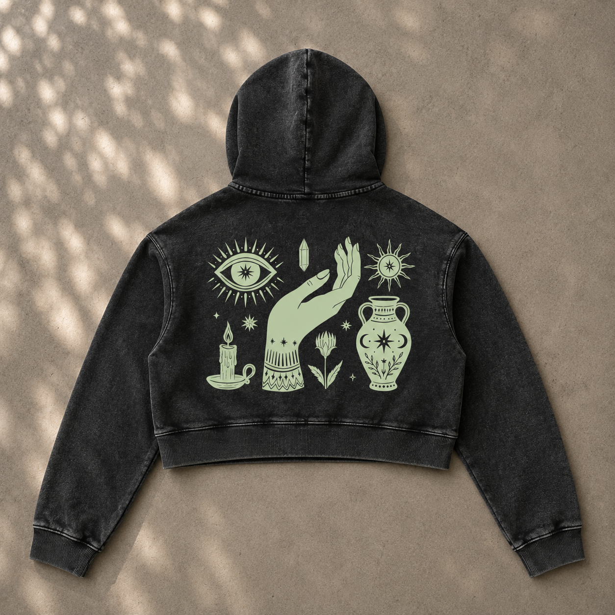 A black washed cropped hoodie shown from the back, featuring a large pale green graphic print of a hand, eye, candle, flower, and urn on a textured beige background with dappled shadows.