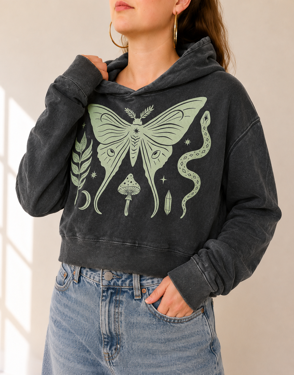 A woman wearing a dark gray washed hoodie with a large light green graphic of a moth, moon, snake, and mushroom, paired with light blue jeans.