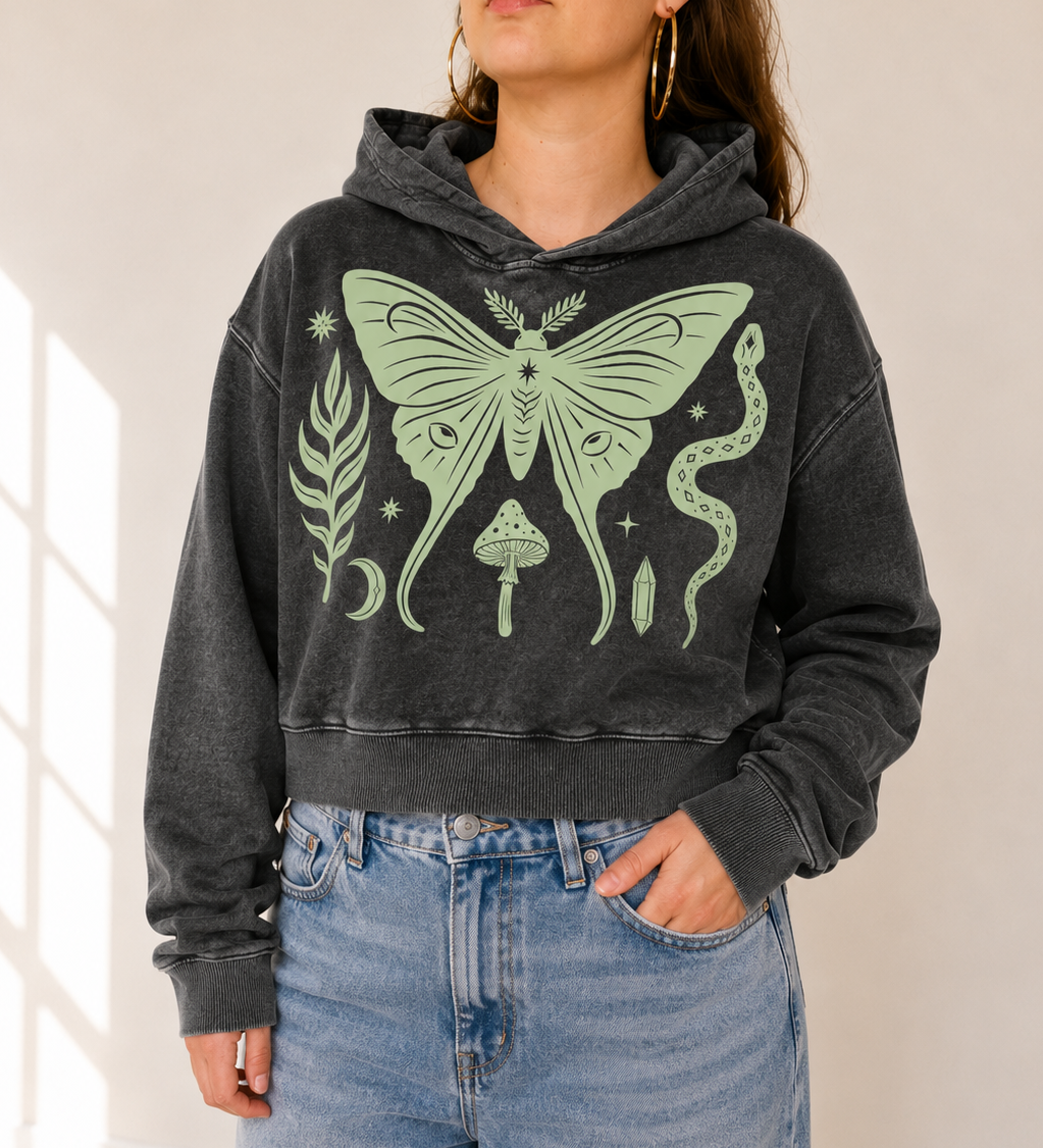 A woman wearing a dark gray washed cropped hoodie with a large light green witch-style graphic on the front, paired with light blue jeans, standing in a brightly lit room.