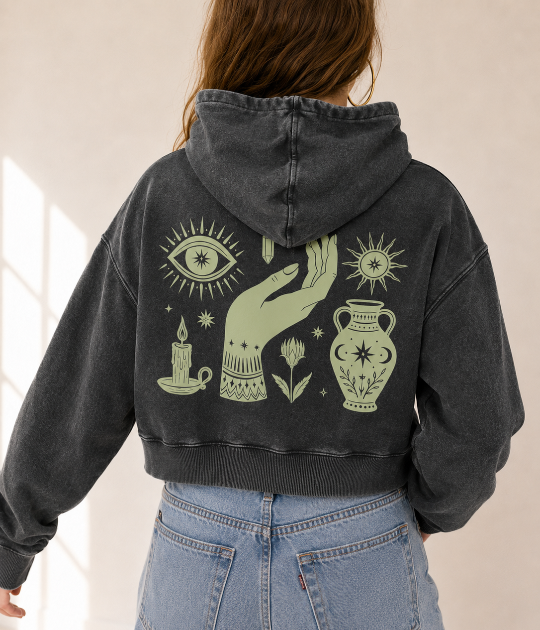 A person viewed from behind wearing a faded charcoal gray cropped hoodie with a large sage green occult-themed graphic on the back, paired with light blue jeans.