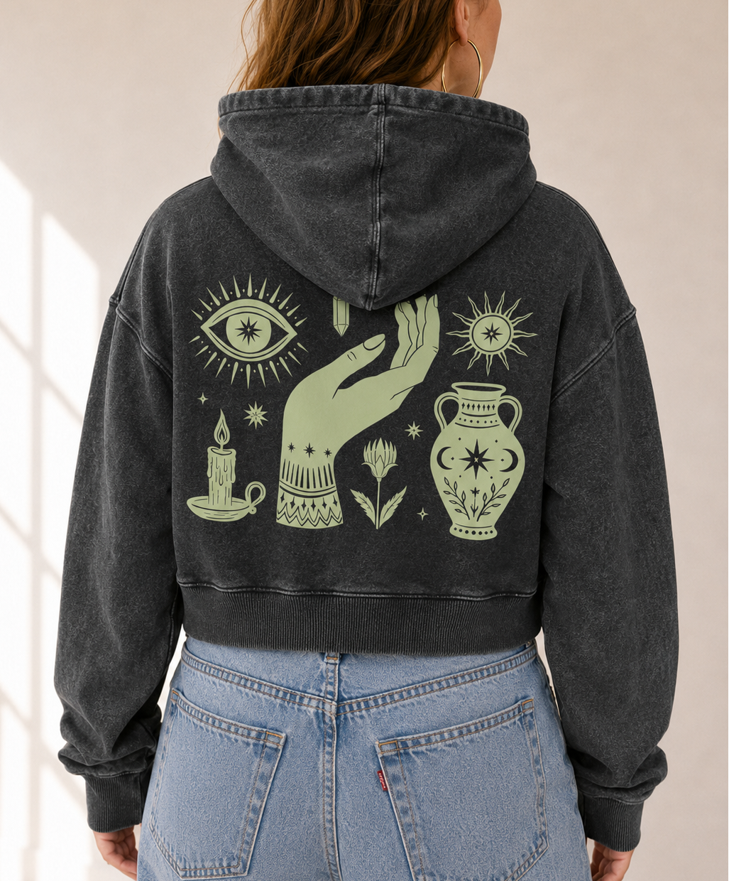 A person viewed from behind wearing a faded black cropped hoodie with a large sage green occult-themed graphic on the back, paired with light blue jeans in a bright indoor setting.