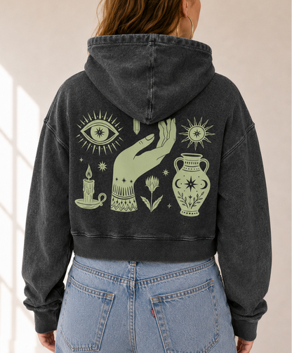 A person viewed from behind wearing a faded black cropped hoodie with a large sage green occult-themed graphic on the back, paired with light blue jeans in a bright indoor setting.