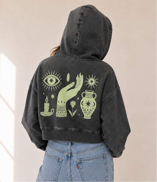 A person shown from the back wearing a faded black cropped hoodie with a large light green occult-style graphic on the back, paired with light blue jeans in a bright indoor room.