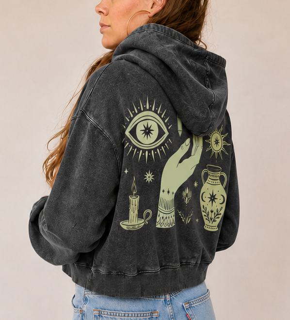 A woman wearing a faded black hooded sweatshirt with a large green and yellow occult-style graphic on the back, paired with light-wash wide-leg jeans, standing in a brightly lit room.