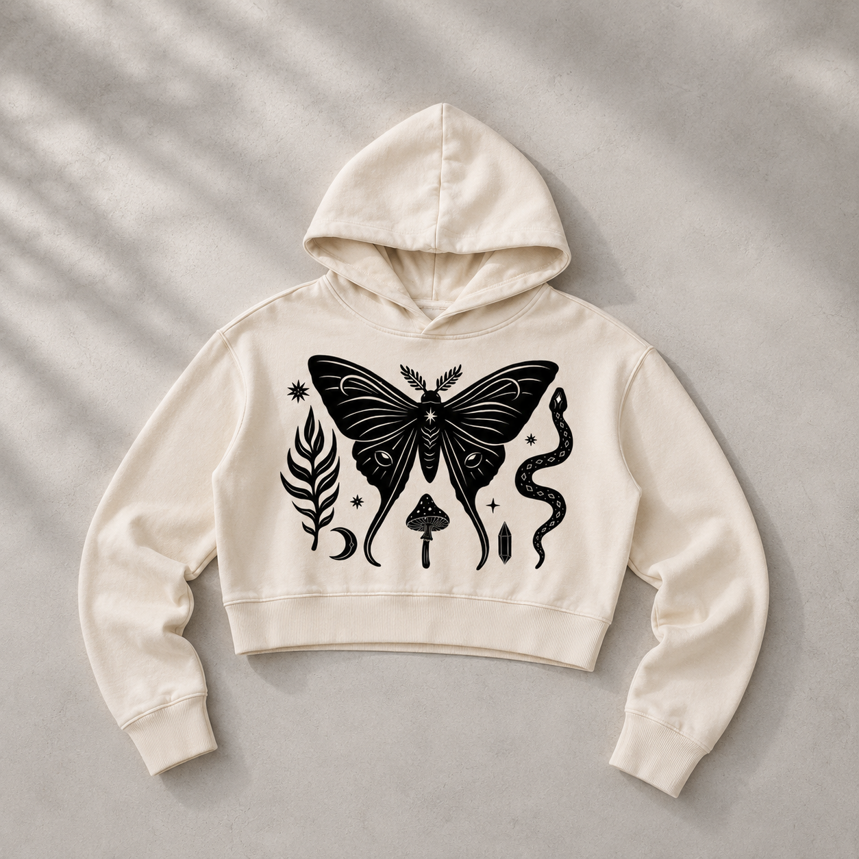 An off-white cropped hoodie with a black graphic print of a moth, a snake, a leaf, and a mushroom, laid flat on a textured gray background with soft leaf shadows.