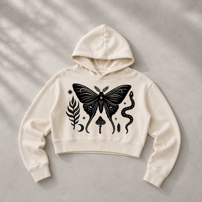 An off-white cropped hoodie with a black graphic print of a moth, a snake, a leaf, and a mushroom, laid flat on a textured gray background with soft leaf shadows.