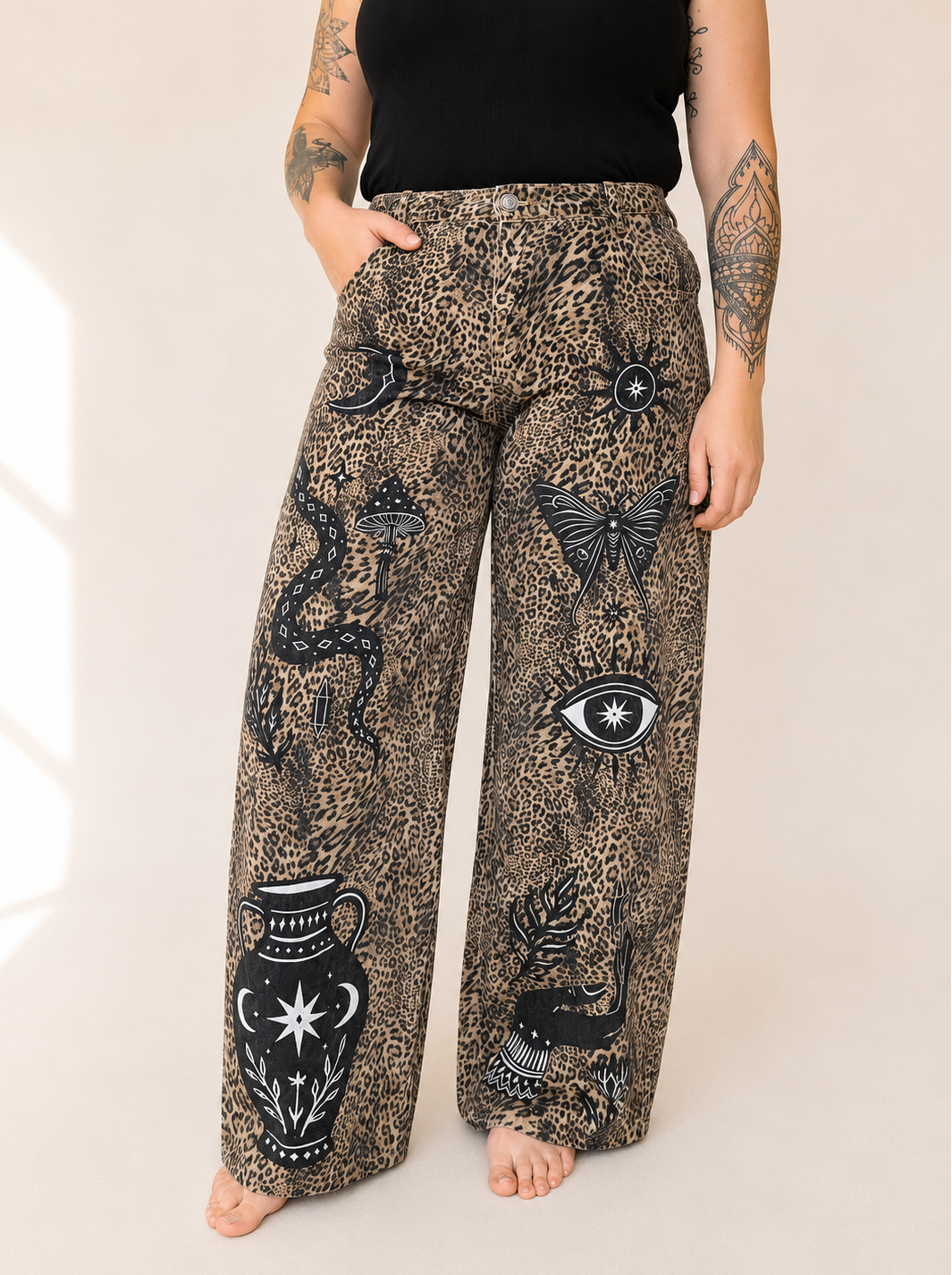 A person wearing loose, wide-leg pants with a leopard-print base and black occult-style illustrations including a snake, eye, butterfly, and vase, paired with a black top against a plain light beige studio background.
