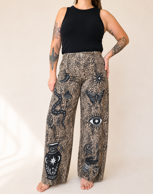 A model wearing wide-leg bohemian pants with a black and brown leopard print and black celestial illustrations, paired with a black sleeveless top, against a warm beige background.
