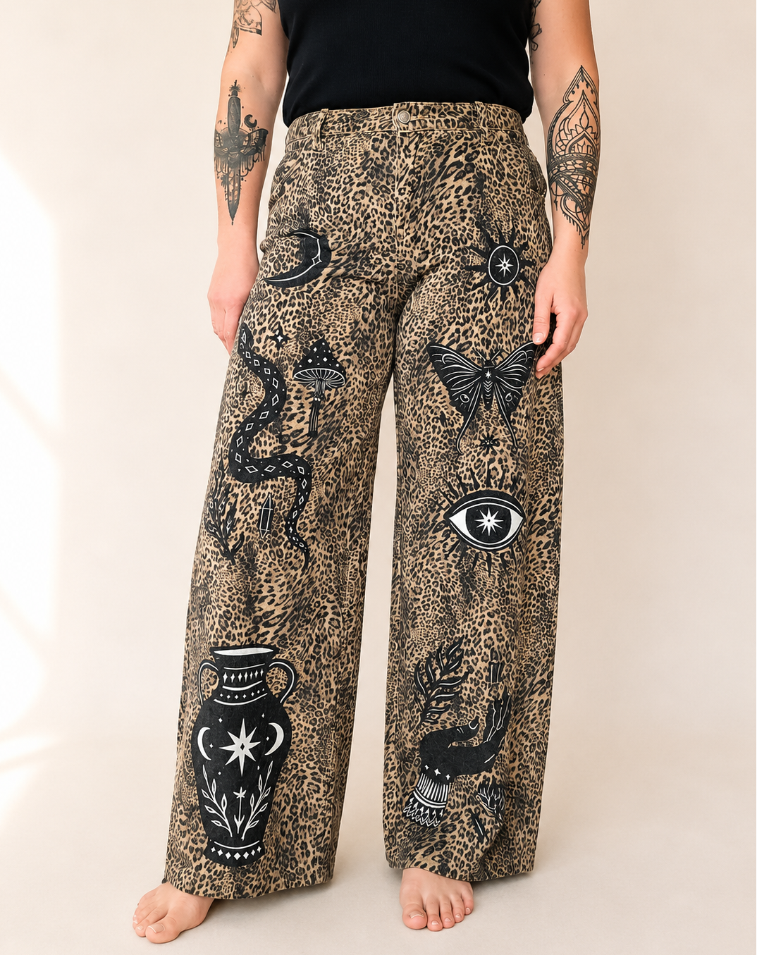 A person wearing wide-leg pants with a beige and brown leopard print base and black occult-style illustrations including a snake, eye, butterfly, vase, and bird, photographed against a plain light beige studio background.