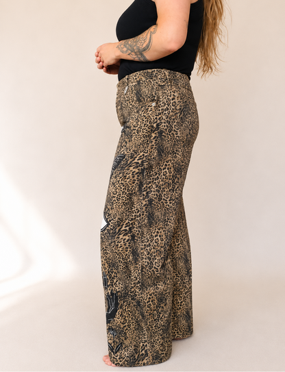 A side view of a model wearing high-waisted, wide-leg pants with a brown and black leopard print pattern, paired with a black sleeveless top against a plain beige studio background.