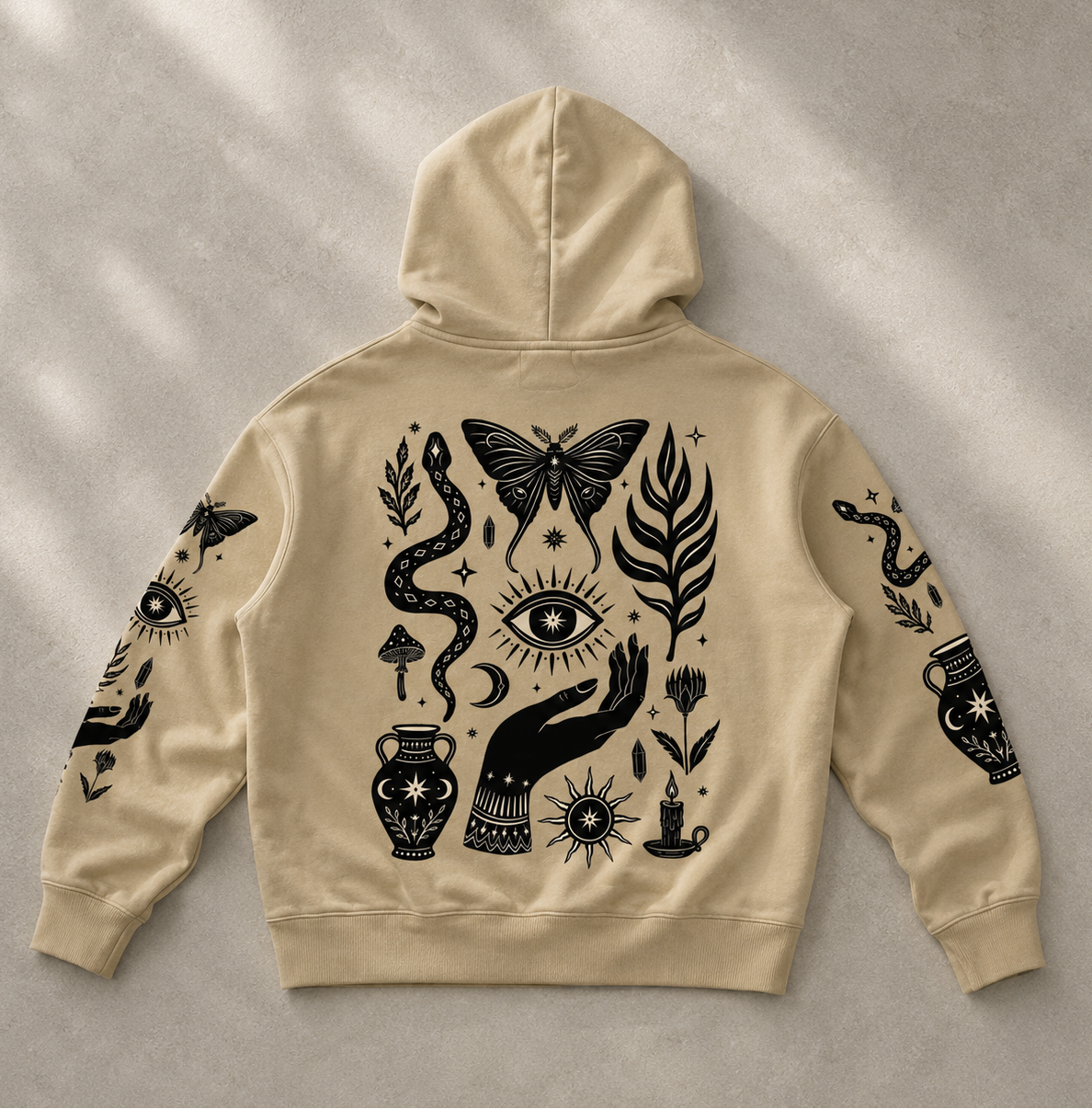 A beige hoodie shown from the back, laid flat on a textured light gray surface with soft diagonal shadows. The hoodie features a large black mystical graphic print including a butterfly, eye, snake, hand, flowers, and a vase.