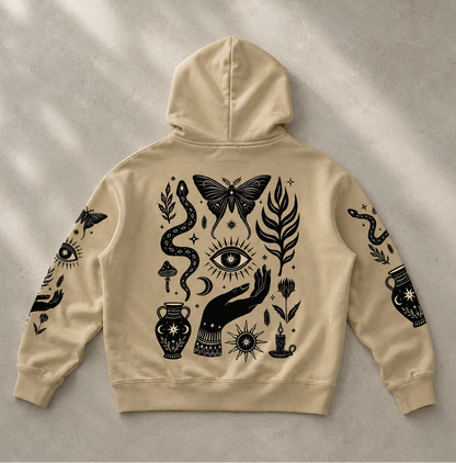 A beige hoodie shown from the back, laid flat on a textured light gray surface with soft diagonal shadows. The hoodie features a large black mystical graphic print including a butterfly, eye, snake, hand, flowers, and a vase.