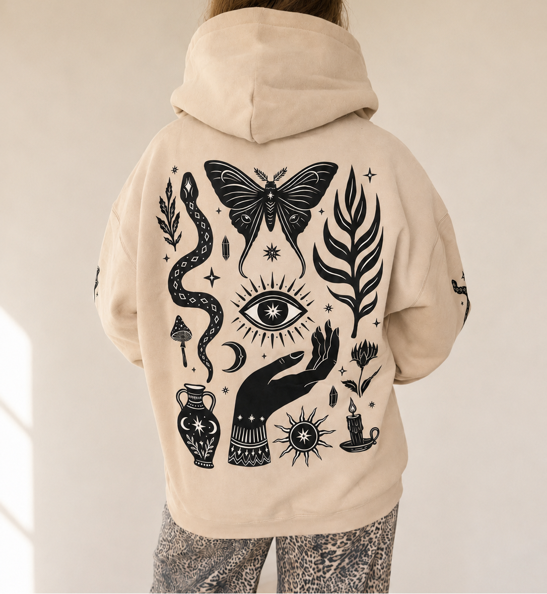 A person shown from behind wearing a beige hoodie with a large black occult-style graphic print on the back, paired with patterned gray pants, in a brightly lit indoor room.