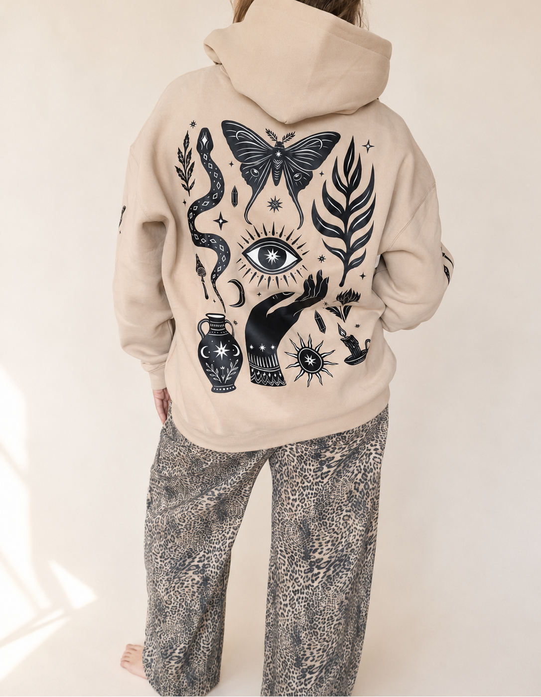 A person shown from the back wearing a beige hoodie with a large black occult-style graphic print featuring a butterfly, eye, snake, hand, and vase, paired with gray patterned pants against a light neutral background.