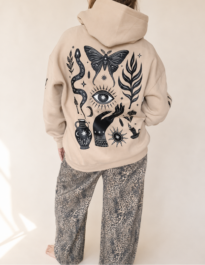 A person shown from the back wearing a beige hoodie with a large black occult-style graphic print featuring a butterfly, eye, snake, hand, and vase, paired with gray patterned pants against a light neutral background.