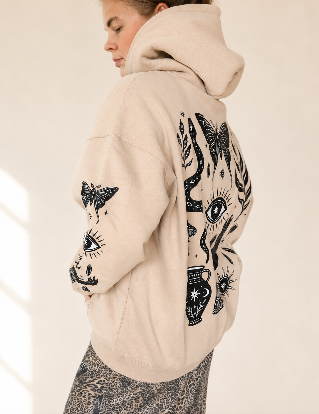 A woman wearing a beige hoodie with black graphic prints of butterflies, eyes, and floral motifs, shown from a side-back angle against a warm off-white background.