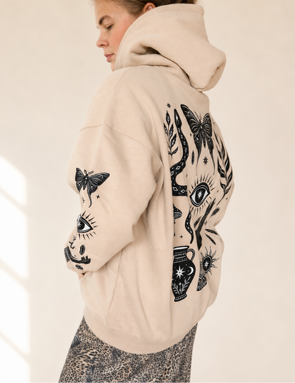 A woman wearing a beige hoodie with black graphic prints of butterflies, eyes, and floral motifs, shown from a side-back angle against a warm off-white background.