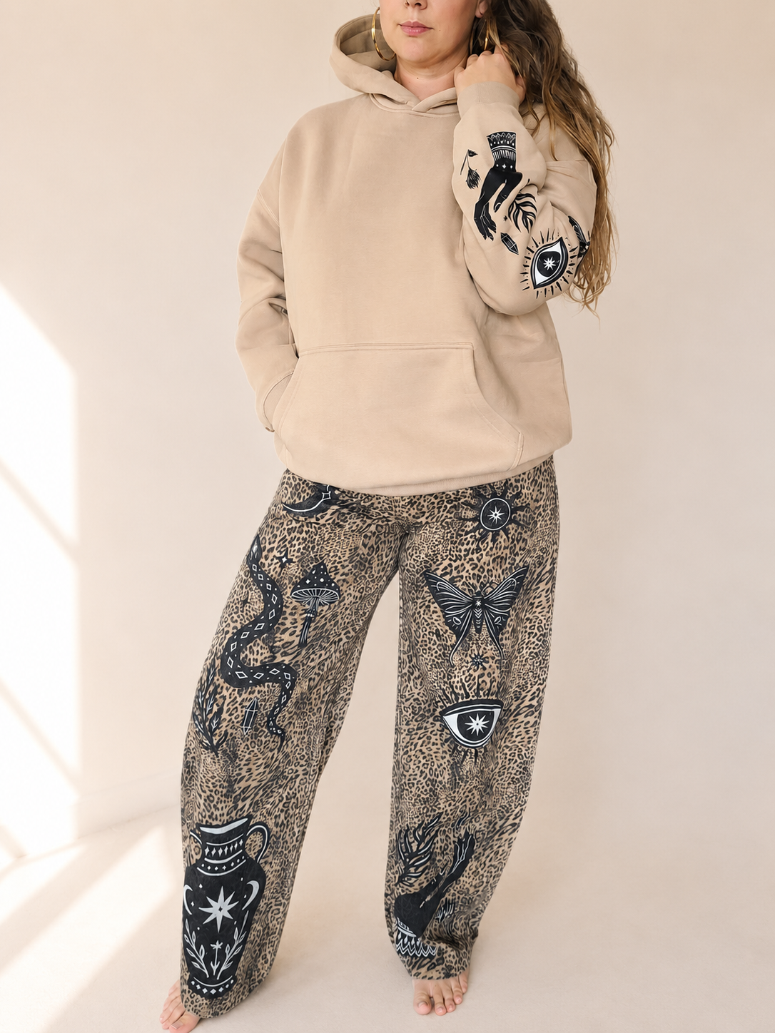 A woman wearing a beige pullover hoodie with black snake and eye graphics on the sleeves, paired with wide-leg patterned pants featuring snakes, butterflies, and eye motifs in black and beige.