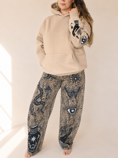 A woman wearing a beige pullover hoodie with black snake and eye graphics on the sleeves, paired with wide-leg patterned pants featuring snakes, butterflies, and eye motifs in black and beige.