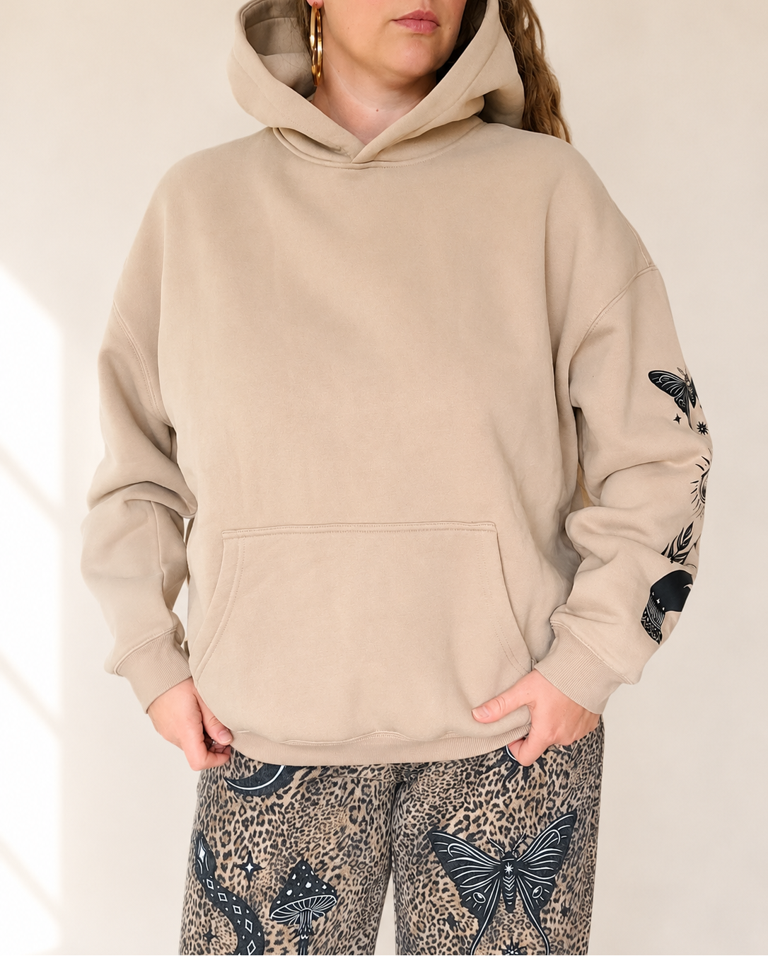 A woman wearing a beige pullover hoodie with black butterfly and snake prints on the sleeves, paired with patterned leopard-print pants, against a warm off-white background.