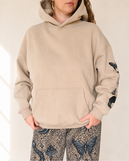 A woman wearing a beige pullover hoodie with black butterfly and snake prints on the sleeves, paired with patterned leopard-print pants, against a warm off-white background.