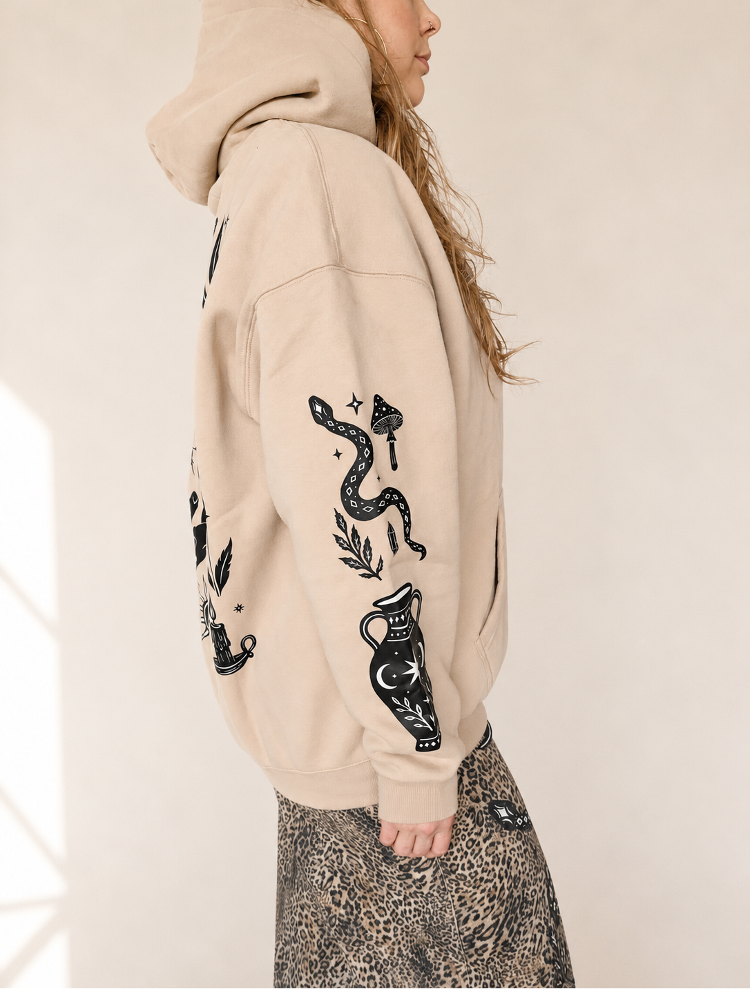 A woman wearing a beige oversized hoodie with black snake and vase graphics on the sleeves, shown from the side against a light neutral background.