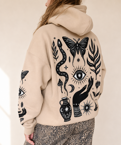 A person shown from the back wearing a beige hoodie with a large black occult-style graphic print featuring a butterfly, snake, eye, hand, vase, and stars. The person is also wearing leopard-print pants against a plain light beige background.