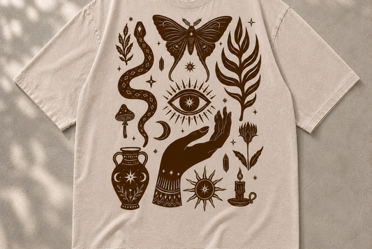 A beige short-sleeve t-shirt with a dark brown bohemian-style graphic print featuring a snake, butterfly, eye, hand, candle, and other mystical symbols, laid flat on a textured gray background with soft leaf shadows.