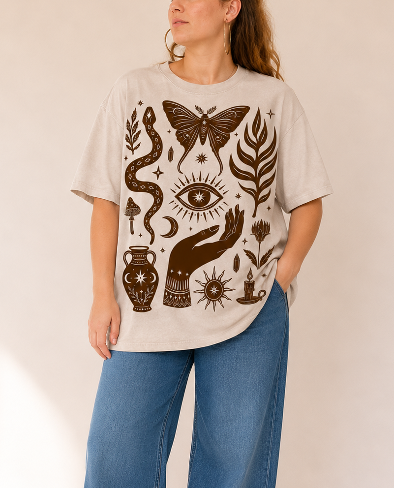 A woman wearing a cream-colored oversized t-shirt with a dark brown bohemian-style graphic print featuring a snake, butterfly, eye, and hand, paired with wide-leg blue jeans, standing against a warm beige background.