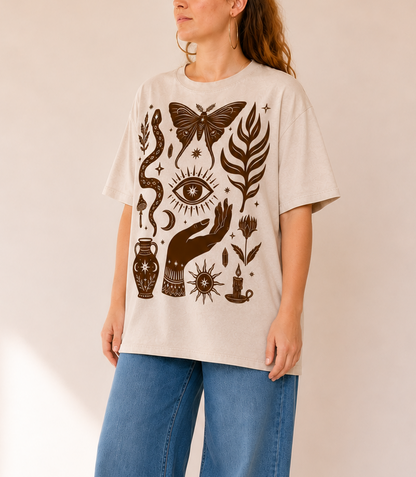 A woman wearing a cream-colored oversized t-shirt with a dark brown bohemian-style graphic print of a butterfly, eye, snake, and hand, paired with wide-leg blue jeans, standing against a warm beige background.