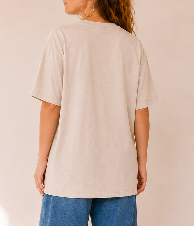 A woman shown from the back wearing an oversized light beige t-shirt and wide-leg blue jeans, standing against a warm beige wall with soft window shadow patterns.