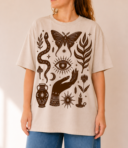 A woman wearing a light beige oversized T-shirt with a dark brown witch-themed graphic print featuring a snake, butterfly, eye, hand, candle, and other mystical symbols, paired with blue jeans against a warm beige background.