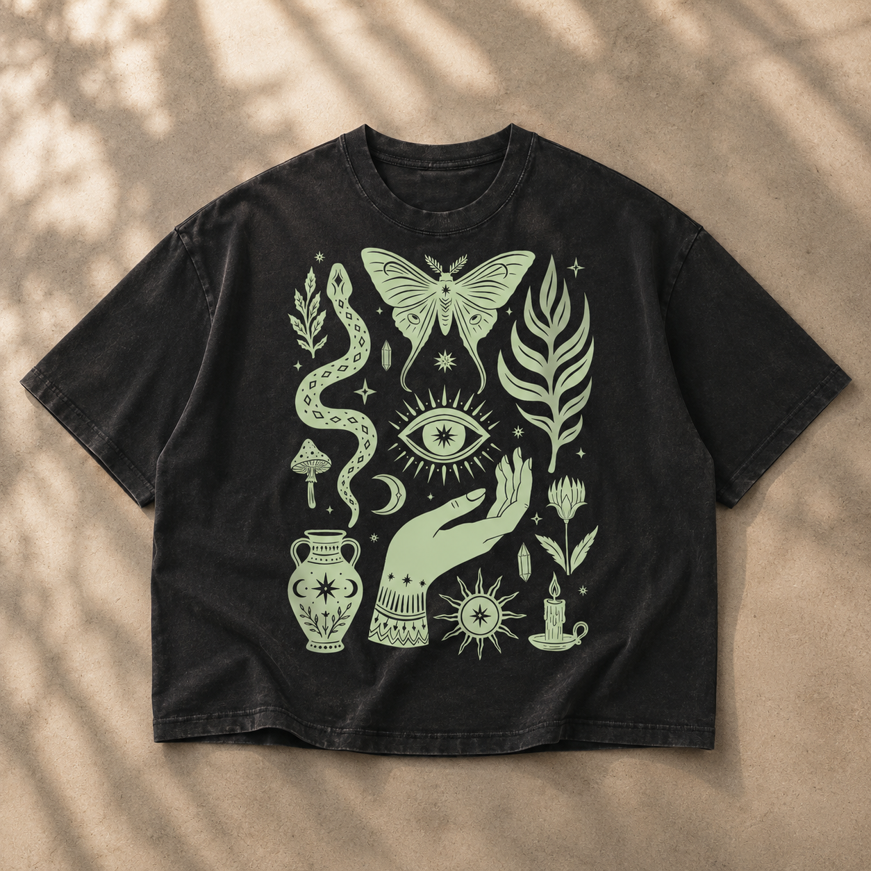 A black washed short-sleeve t-shirt with a light sage green bohemian-style graphic print, laid flat on a textured beige surface with soft leaf shadows.
