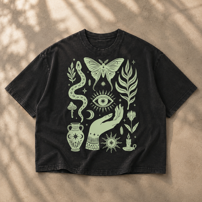 A black washed short-sleeve t-shirt with a light sage green bohemian-style graphic print, laid flat on a textured beige surface with soft leaf shadows.