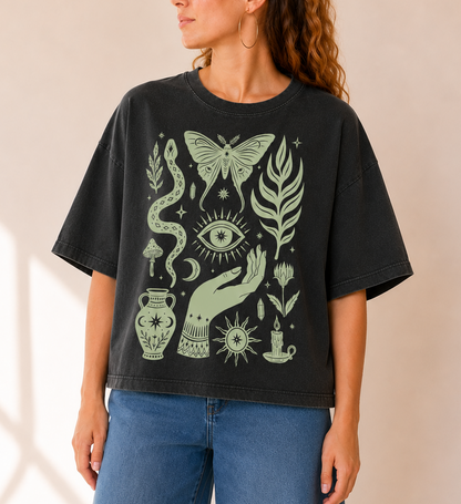 A woman wearing a black graphic t-shirt with a sage green mystical design featuring a butterfly, eye, snake, and hand, paired with blue jeans against a warm beige background.