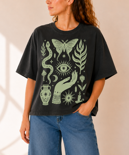 A woman wearing a black graphic t-shirt with a sage green mystical design featuring a snake, eye, hand, and butterfly, paired with blue jeans against a warm beige background.