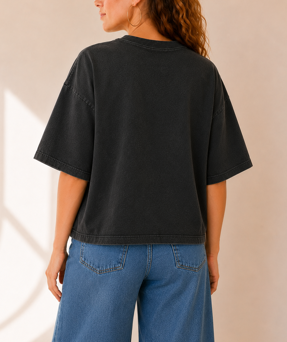 A woman shown from the back wearing an oversized faded black t-shirt and blue jeans, standing against a warm beige wall with soft window shadows.