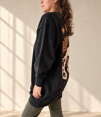 A woman wearing a washed black oversized sweatshirt with a large graphic print on the back, standing in profile against a textured beige wall with soft window shadows.