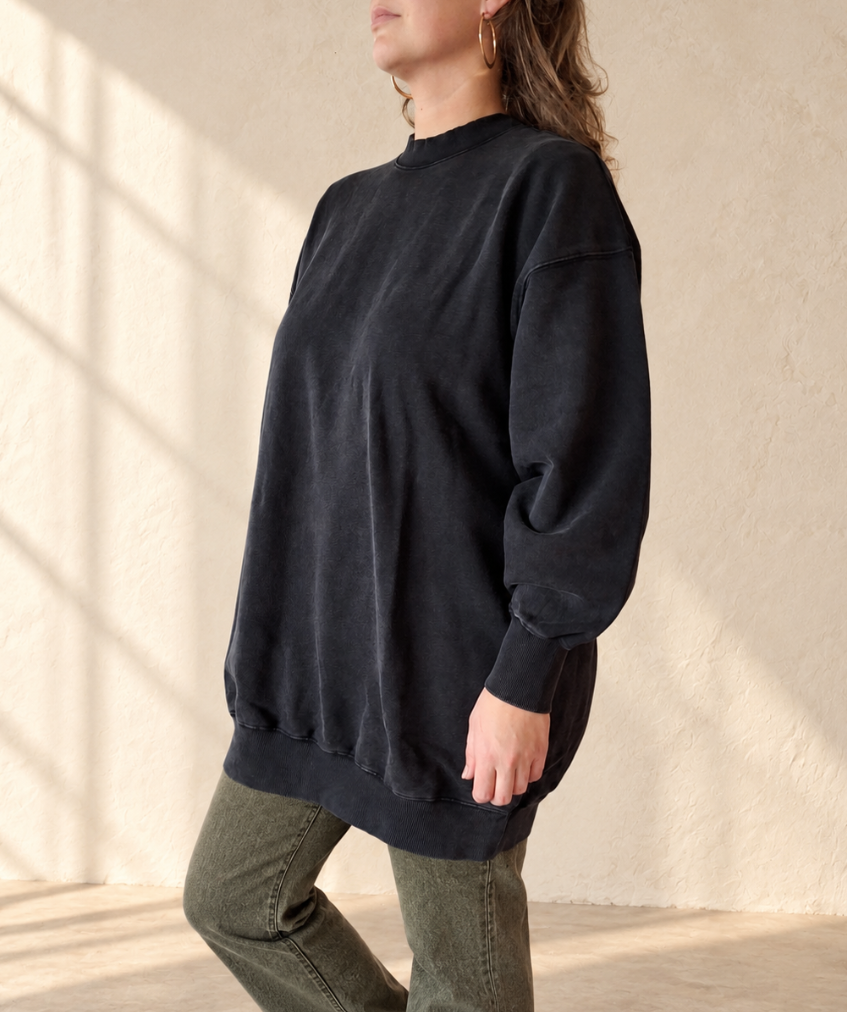 A woman wearing an oversized black crewneck sweatshirt and olive green pants, standing in a room with textured beige walls and soft window shadows.
