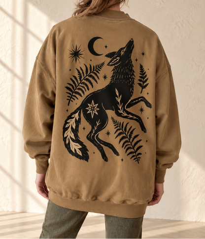 A person shown from the back wearing a tan oversized sweatshirt with a large black graphic of a howling wolf, ferns, stars, and a crescent moon. The photo is taken in a softly lit indoor setting with a textured cream wall and floor shadows.