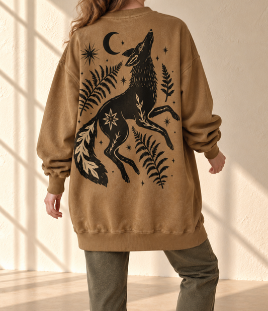 A person shown from the back wearing a tan oversized sweatshirt with a large black graphic of a howling wolf surrounded by ferns, stars, and a crescent moon, paired with olive pants in a sunlit room.