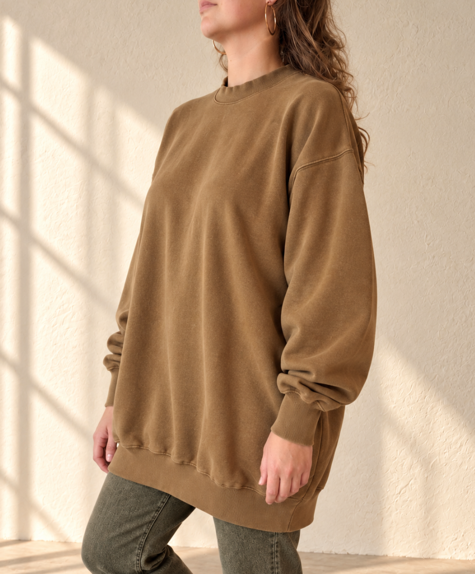 A woman wearing an oversized brown crewneck sweatshirt and olive green jeans, standing against a textured beige wall with soft window shadow patterns.